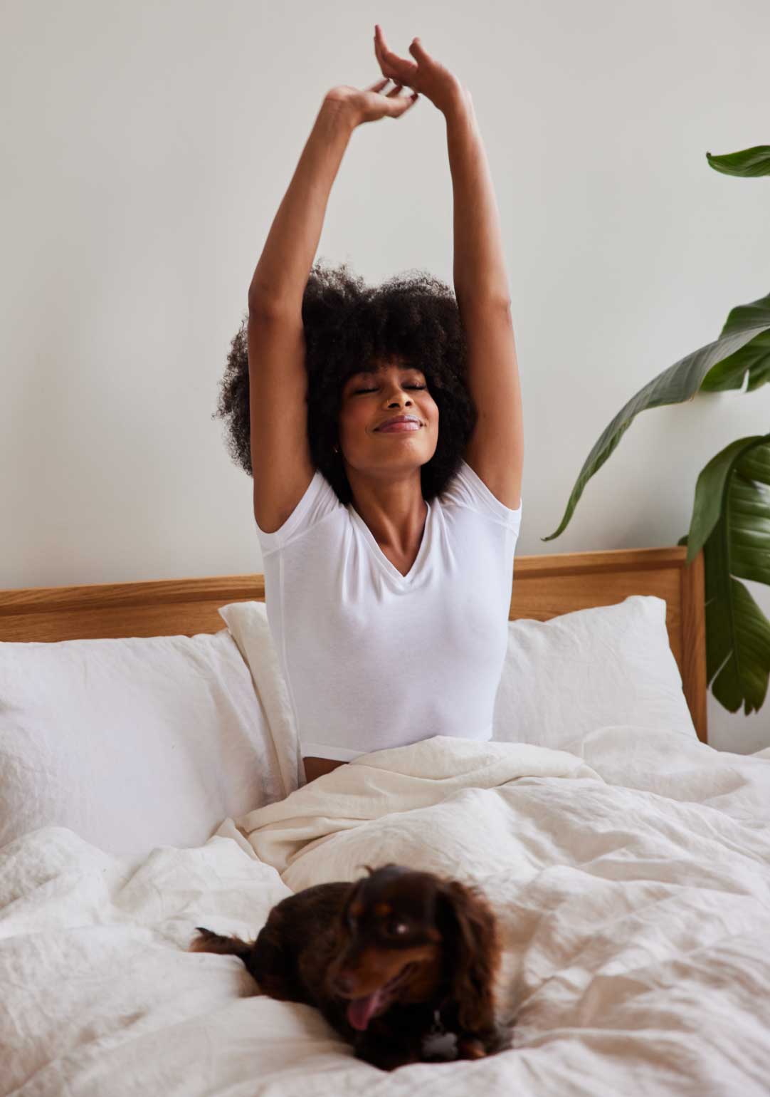 a woman sitting in bed smiling with her arms stretching overhead while her dog lays next to her