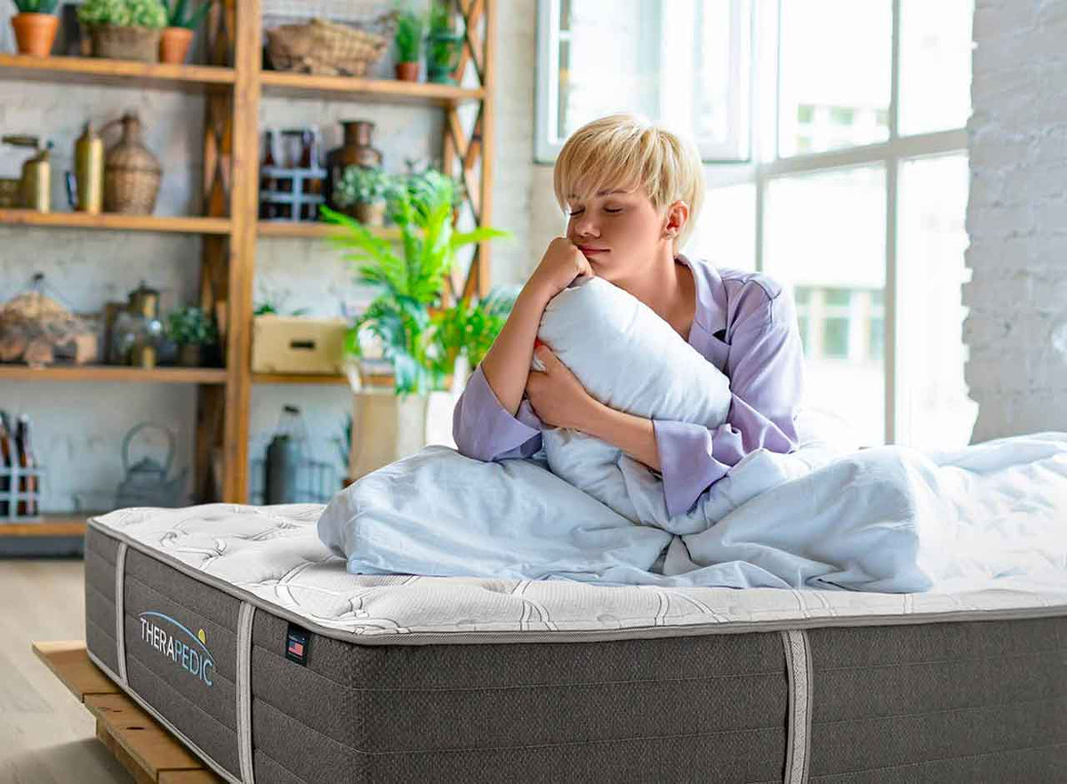 Woman sitting on a Therapedic mattress in a bright room setting, hugging a pillow.