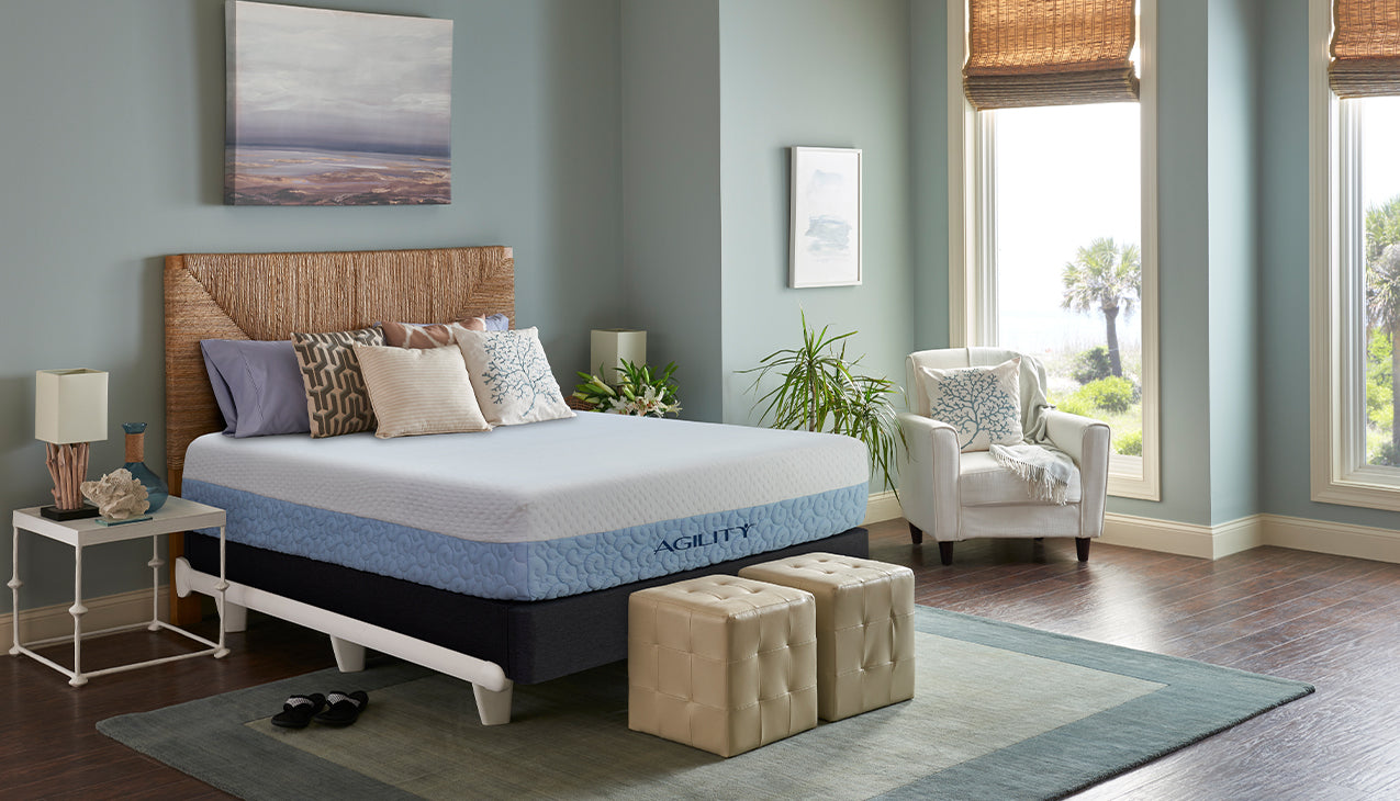 The agility mattress and foundation in a bright bedroom settings on top of a bed frame with a headboard and two ottomans at the foot of the bed.